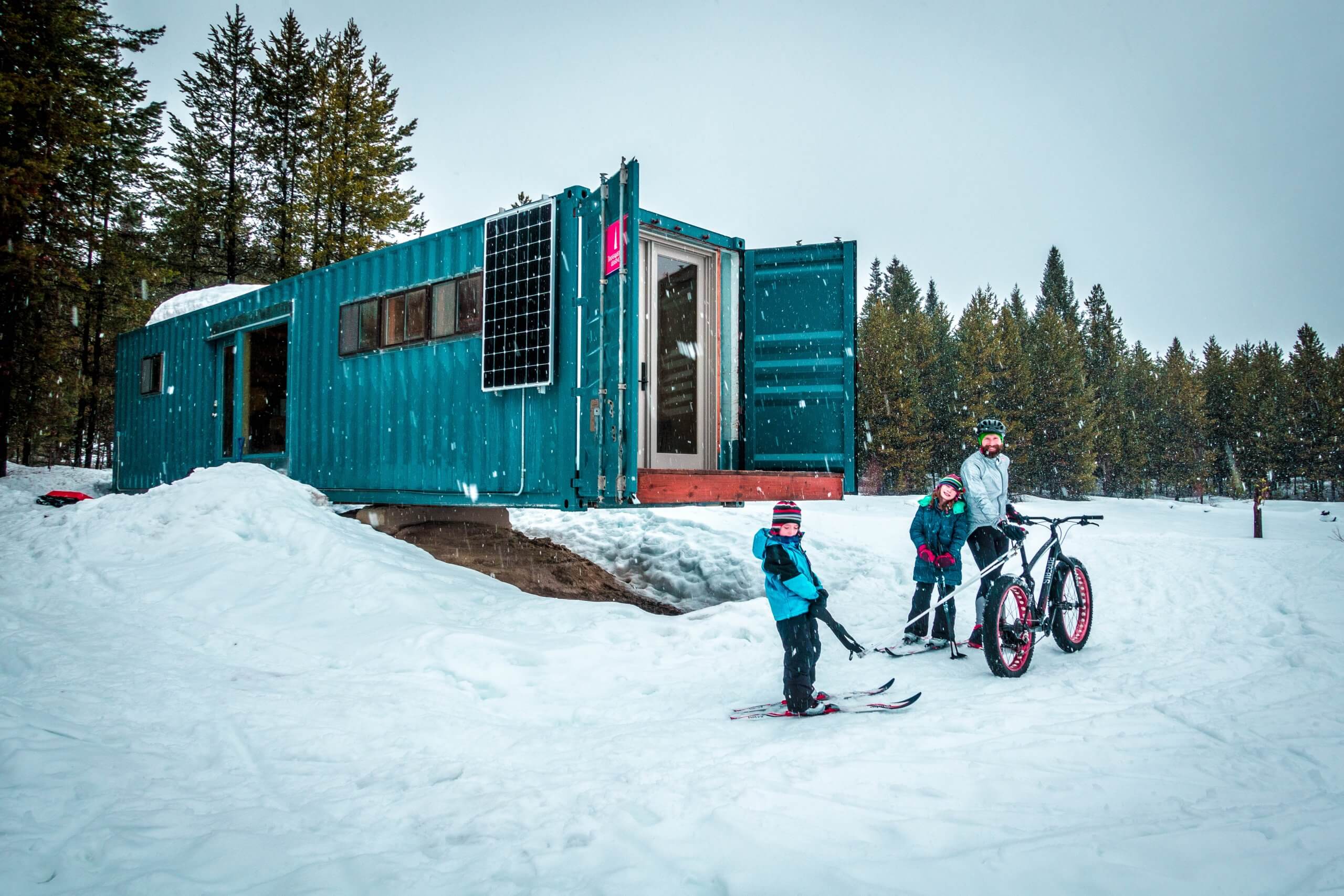 A man and two children with a fat bike and ski gear standing in front of the cargo container cabin at Jug Mountain Ranch, and a forest of trees in the background.