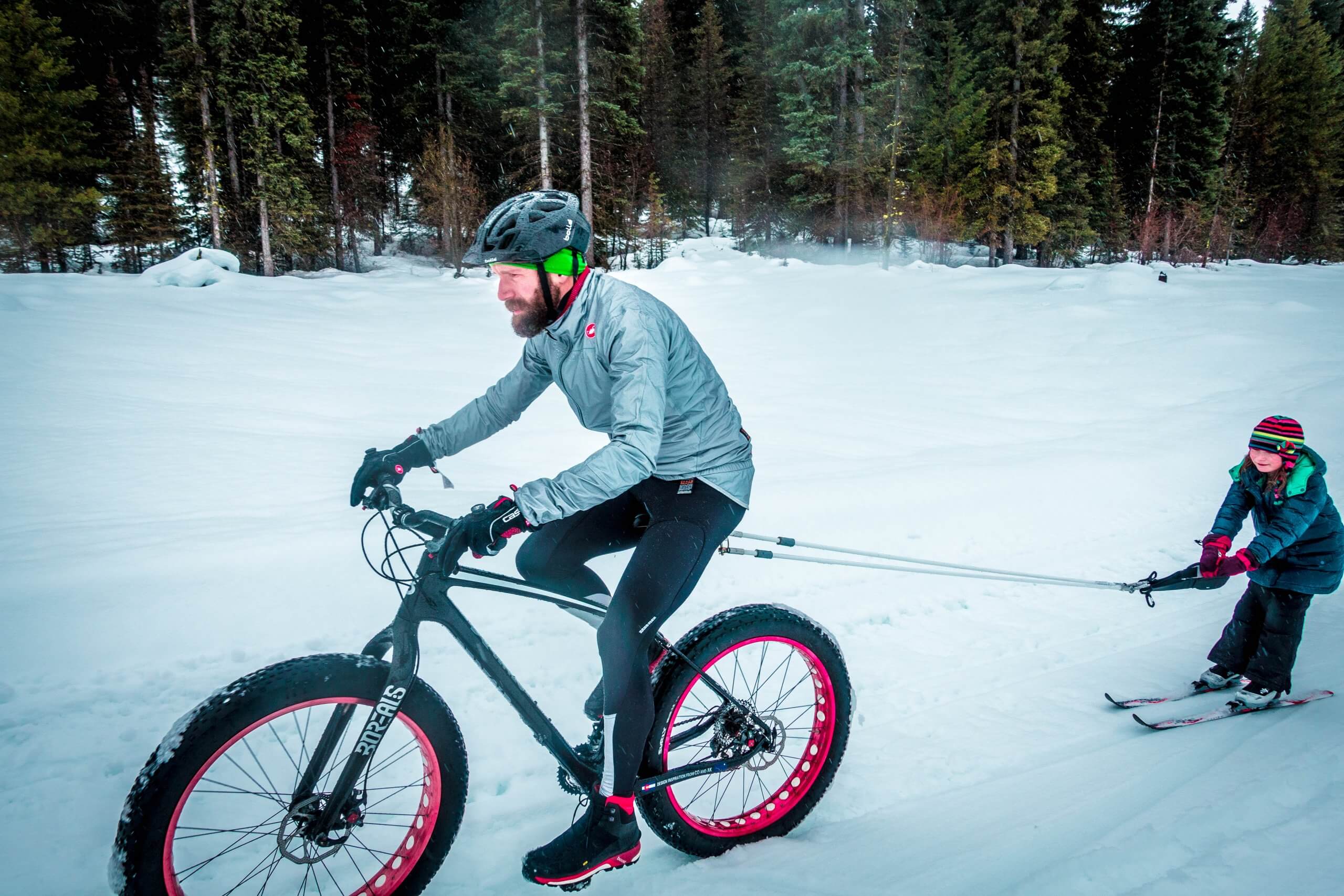 A man on fat bike pulling a child on skis across an open field of snow surrounded by tall trees at Jug Mountain Ranch.