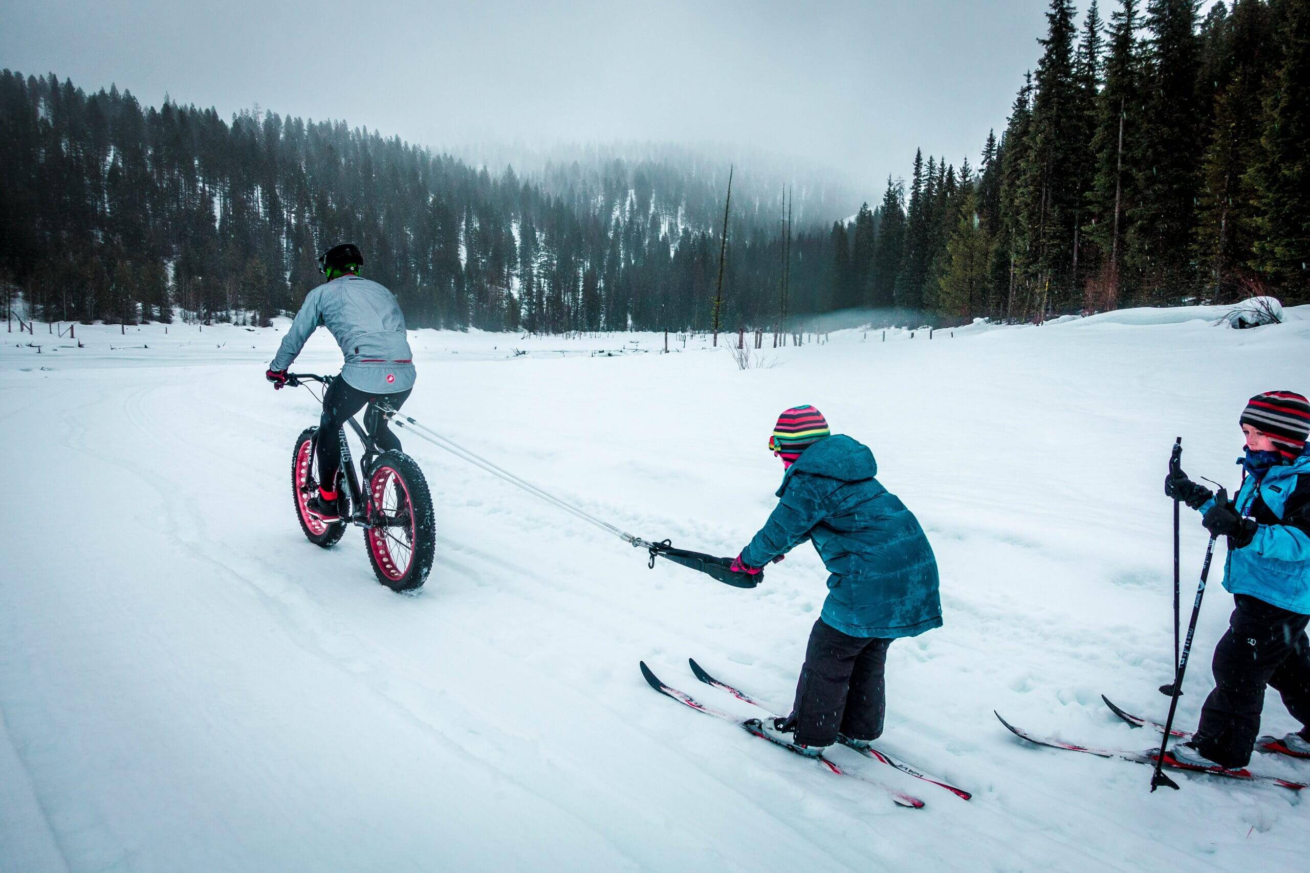 A man pulling a child on skis with fat bike across an open field of snow surrounded by tall trees at Jug Mountain Ranch.