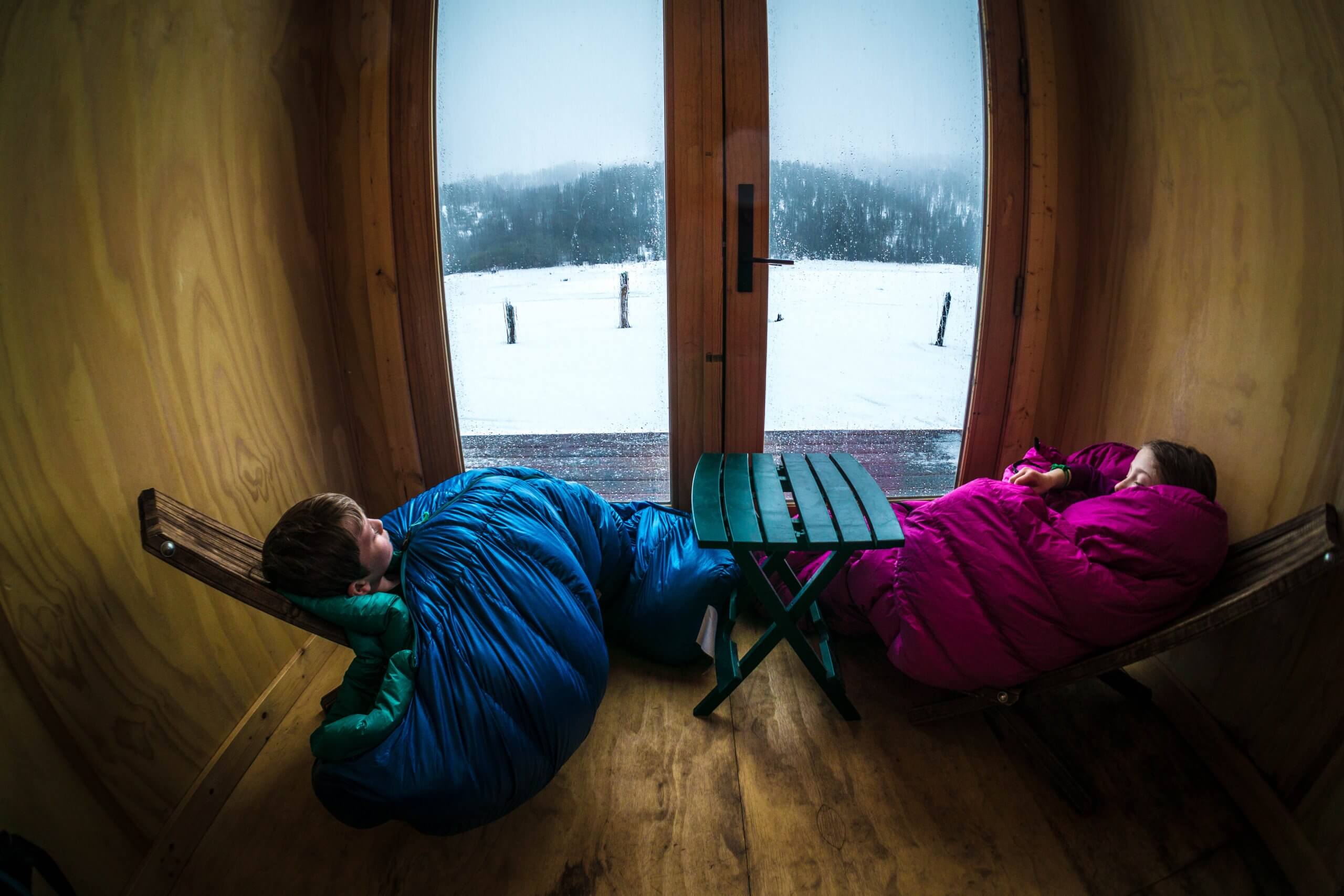 Two kids snuggled up in sleeping bags inside the cargo container cabin at Jug Mountain Ranch, looking out at snow-covered landscape and trees.