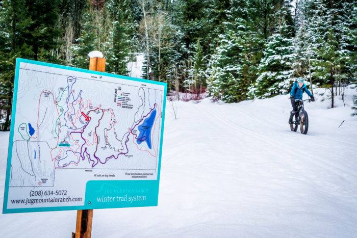 A sign with a map of the Jug Mountain Winter Trail System in the foreground and a person riding a fat bike through a snow-covered forest in the background.