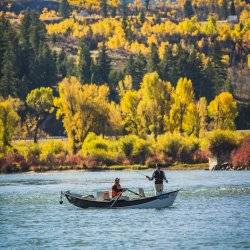 Two men cast lines from a drift boat on a river surrounded by fall colors.