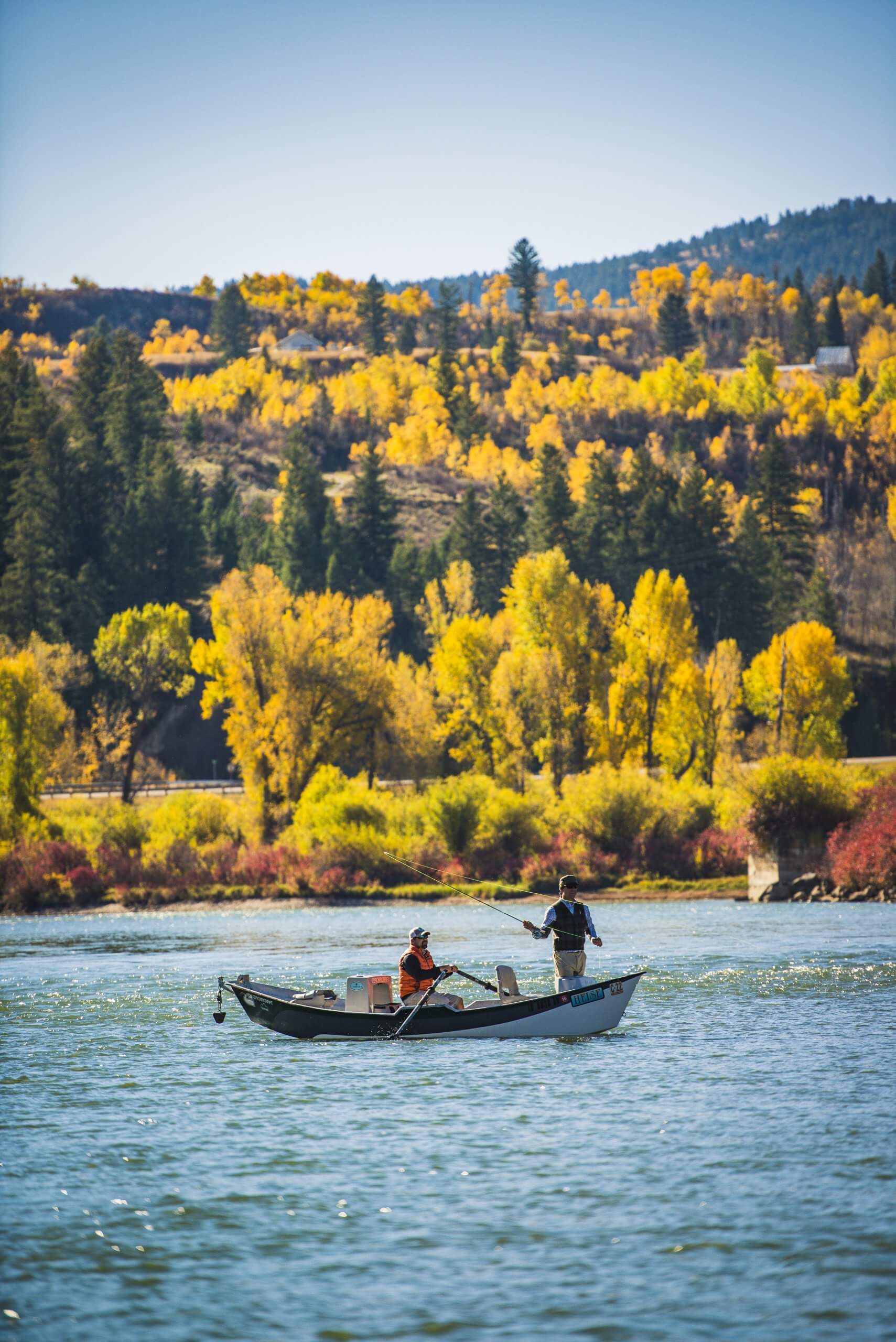 Two men cast lines from a drift boat on a river surrounded by fall colors.