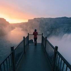 A woman stands at the end of a viewing deck, surrounded by mist from Shoshone Falls at sunset.