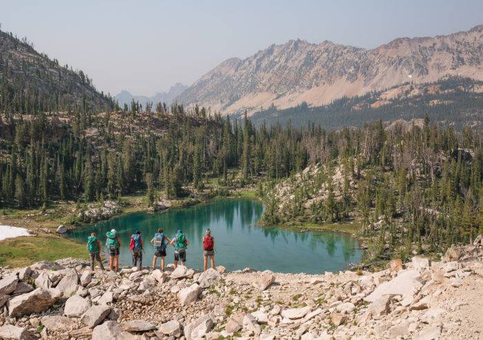 A group of six backpackers standing above an alpine lake with mountains in the background. 