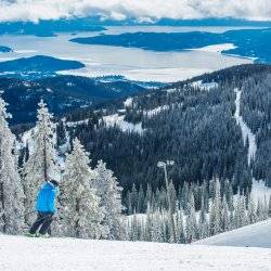 A person in blue coat skiing atop a mountain peak at Schweitzer and a vast forest in the distance.