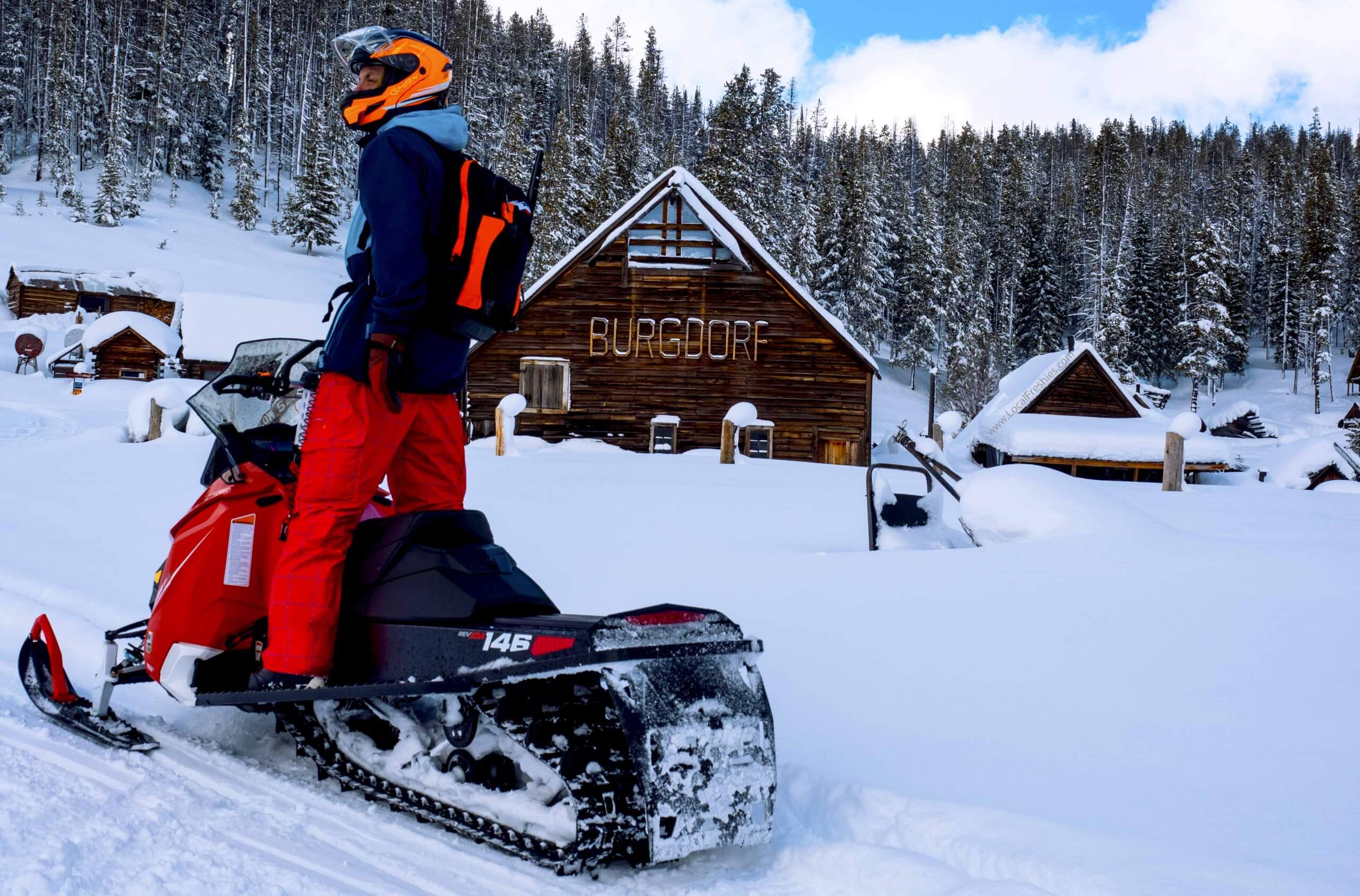 A man in red pants and an orange helmet stands on a snowmobile at Burgdorf Hot Springs.