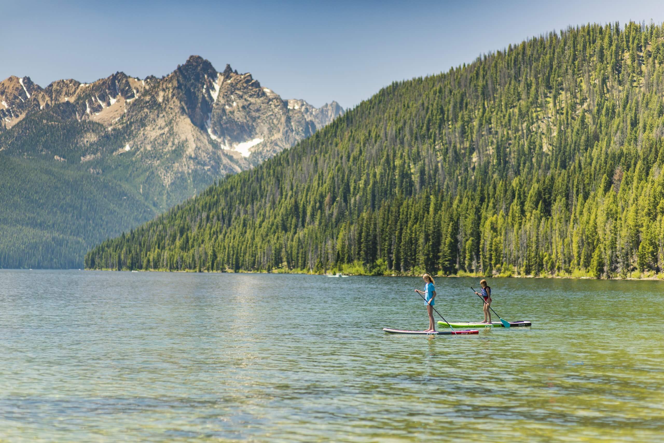 Two people stand up paddleboarding on Redfish Lake and tree-covered mountains in the distance.