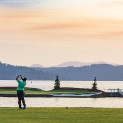 A man swings a golf club on a course near a lake.