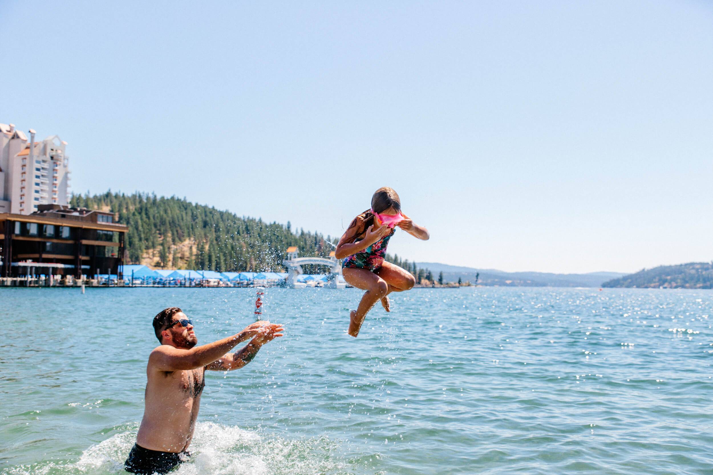 A man tossing his daughter in the water at Lake Coeur d'Alene, with buildings, a marina and a forest in the background.