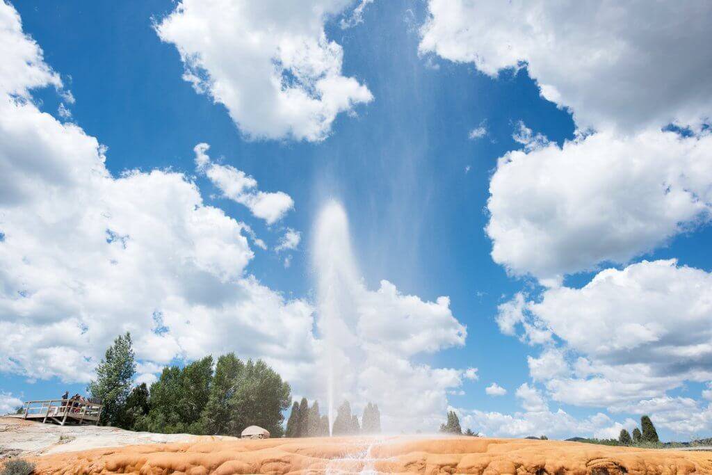 The Soda Springs Geyser erupts against a blue sky with trees in the background.