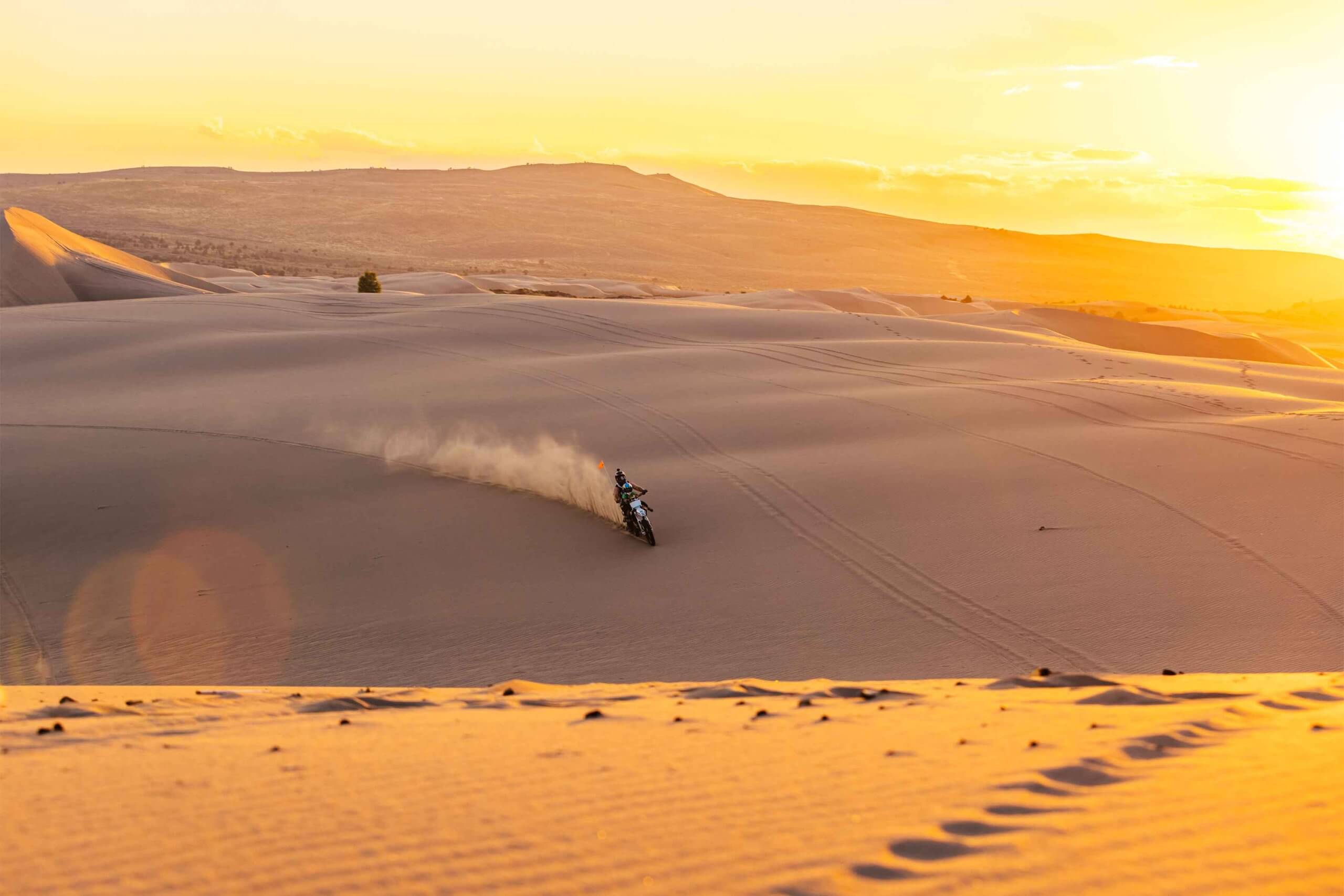 From a distance at sunset, a traveler rides a fat tired bike, leaving behind a trail of dust at the St. Anthony Sand Dunes.