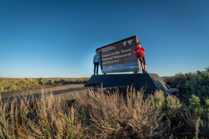 Two people standing next to the Craters of the Moon National Monument & Preserve sign, surrounded by desert brush.