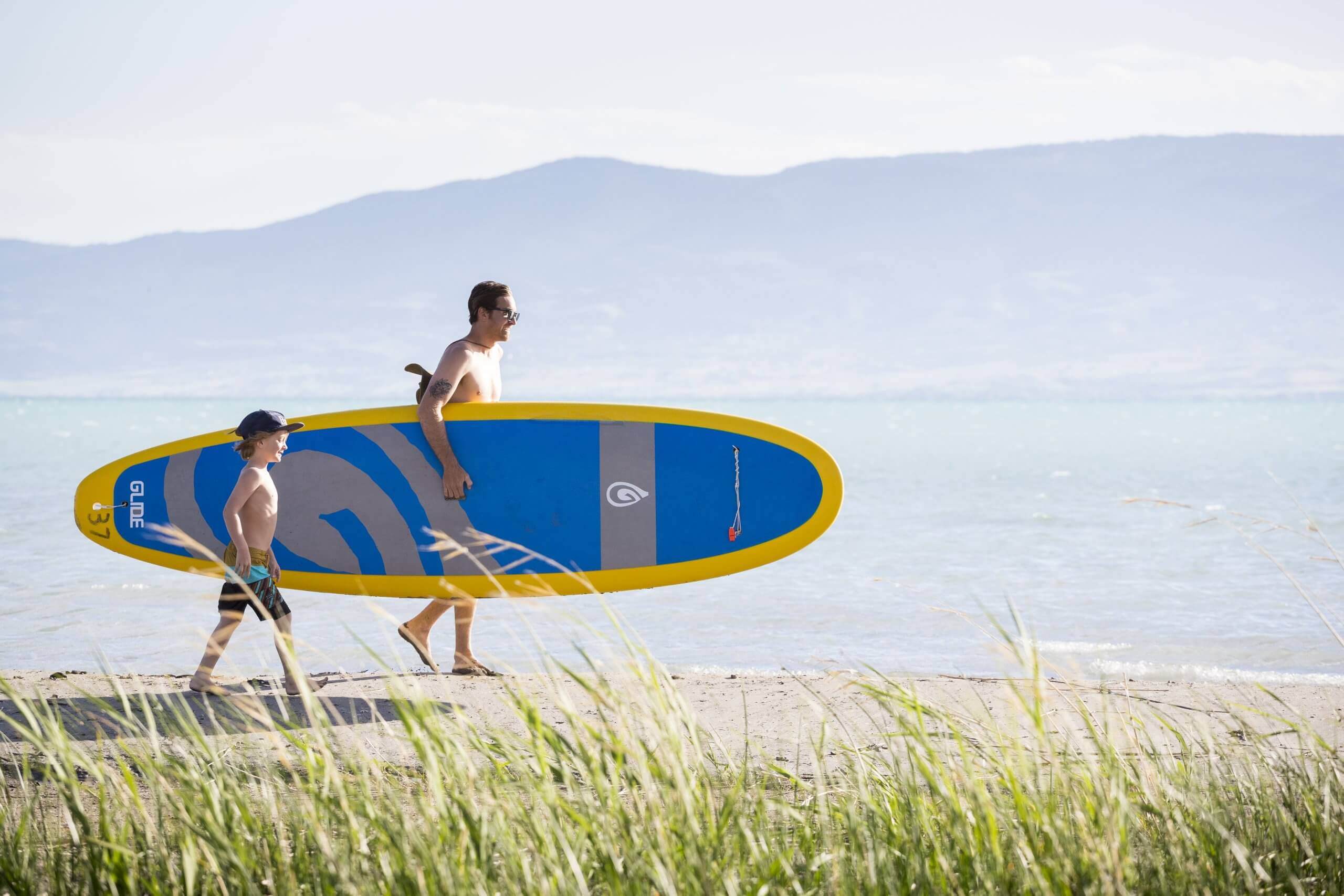 A father and son walking along the beach at Bear Lake, carrying a stand up paddleboard.