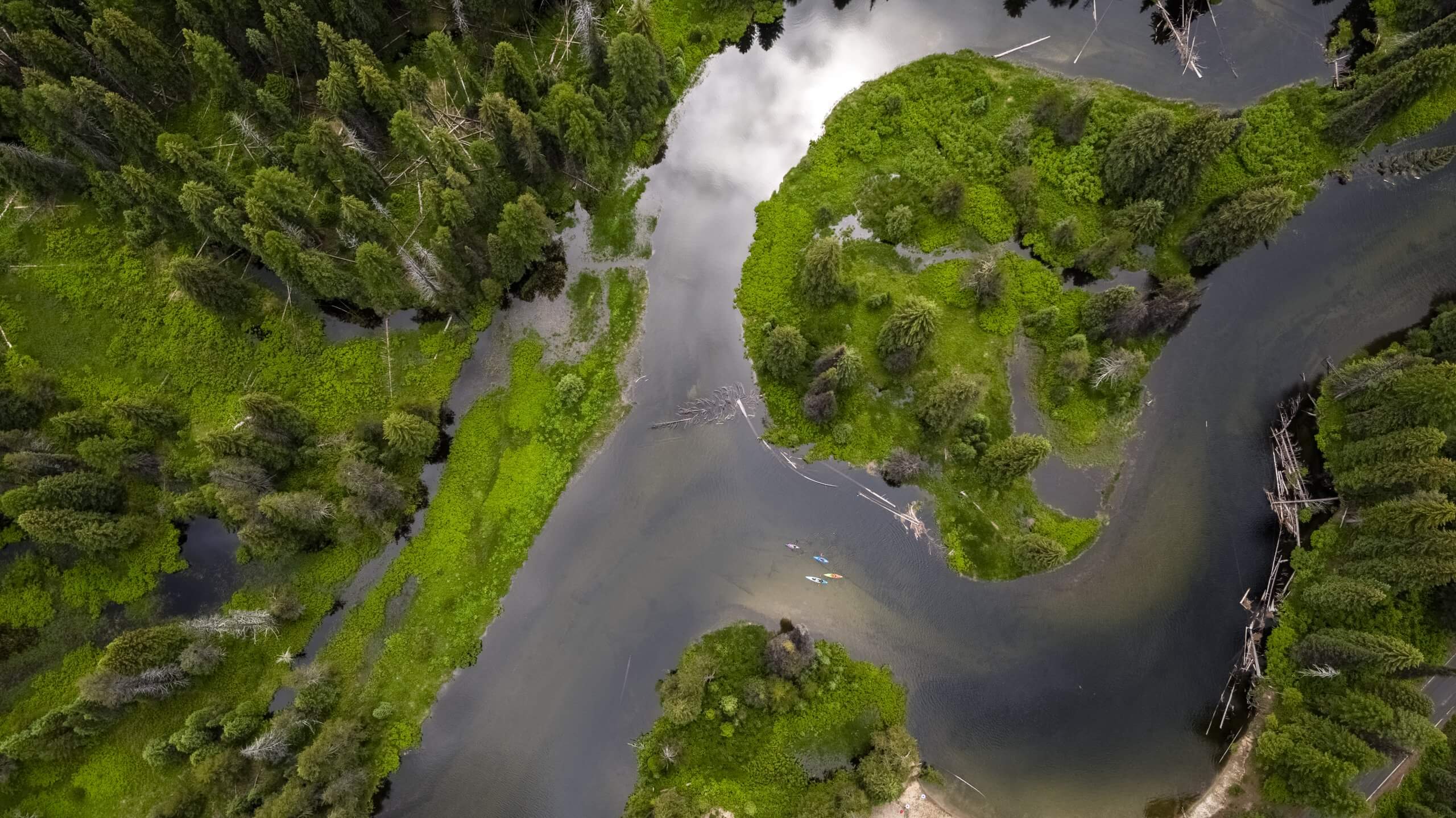 An aerial snapshot picture from above the North Shore Streams at Payette Lake, near McCall, Idaho, where kayakers from down below cross the water, clouds reflected in the lake as lush green trees rise from the distance, captured by a drone.