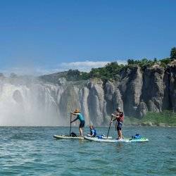 A family of paddleboarders keep a safe distance from the base of the Shoshone Falls as the waterfalls cascade and mist from a distance into the bright waters.