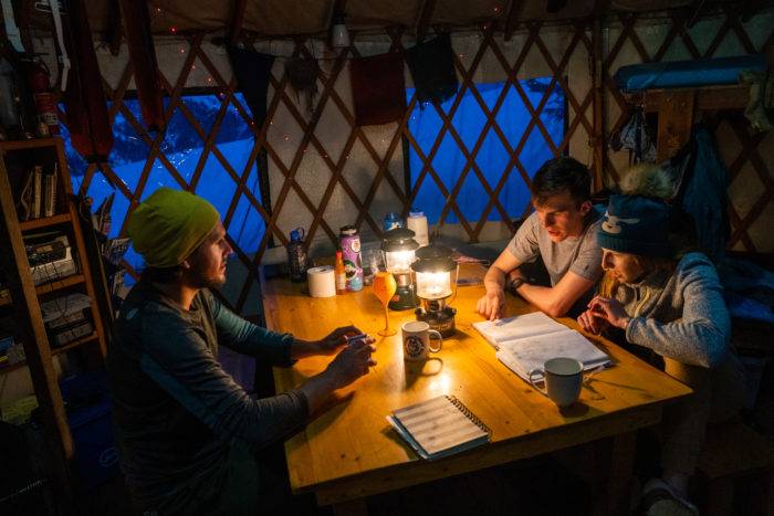 A group of three people sitting at table in a yurt lit by lantern. 