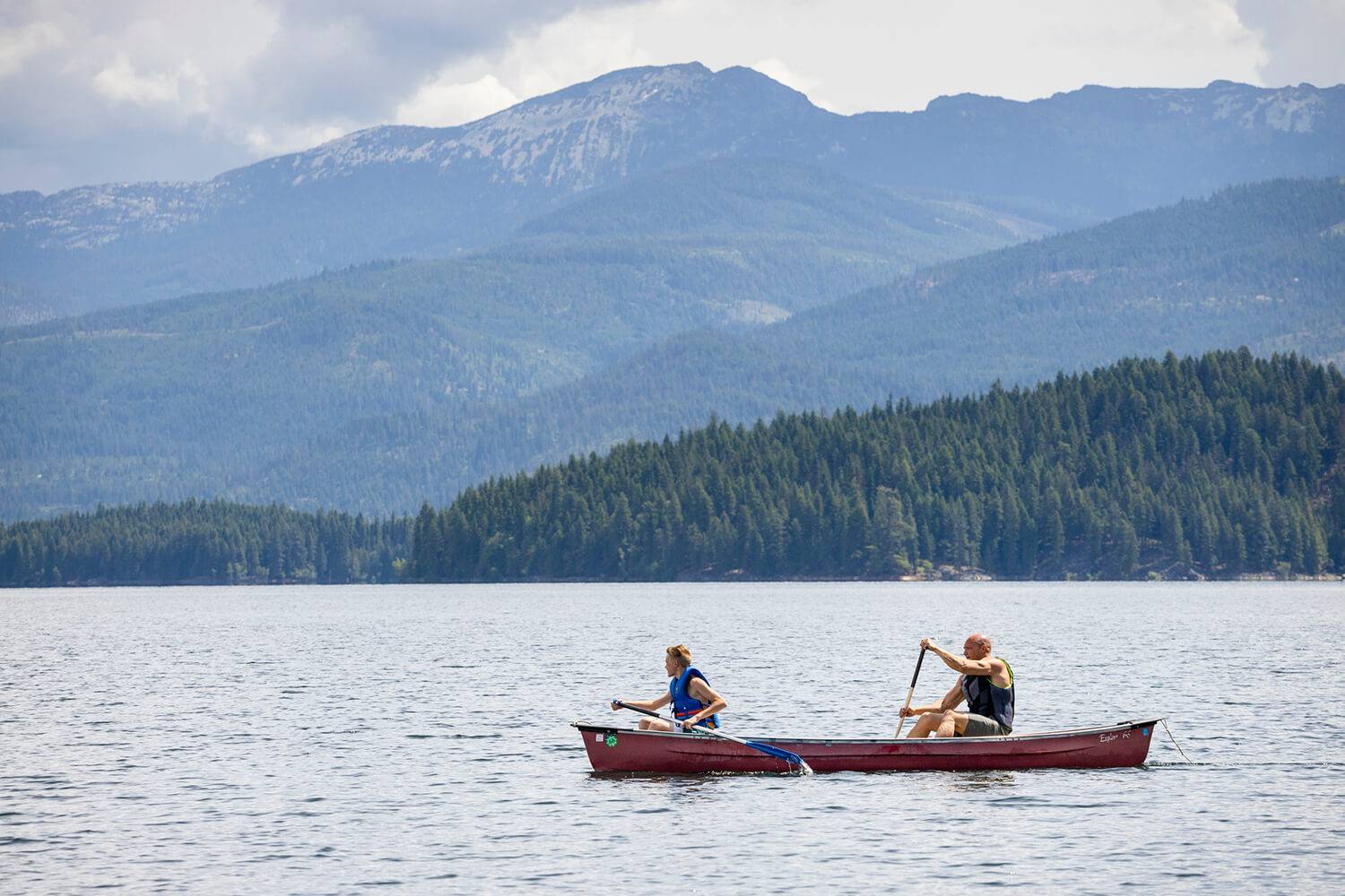 Two people paddling a canoe on Priest Lake with tree-covered mountains in the background.