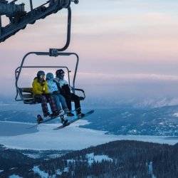 Three people in ski gear sitting on a chair lift during sunrise with mountains and a lake behind them.