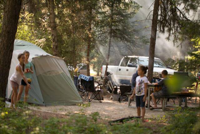 Three children play around their campsite, with tall trees and a sage green colored tent, and white pick up truck in the background.