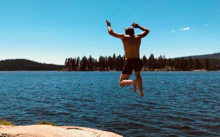 man wearing swim trunks jumping into a lake and a forest of trees in the distance