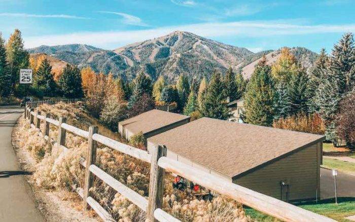 curving road lined by a wooden fence with buildings to one side and trees and mountains in the background