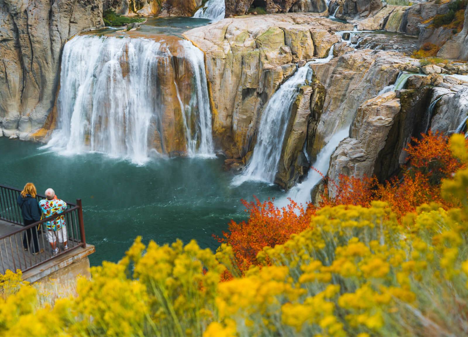 Two people stand at a viewpoint at Shoshone Falls with fall foliage in the foreground.