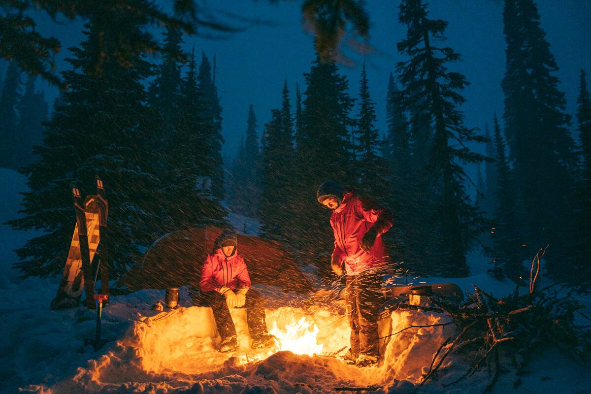 Two people in red jackets gather around a campfire in a snowy forest.