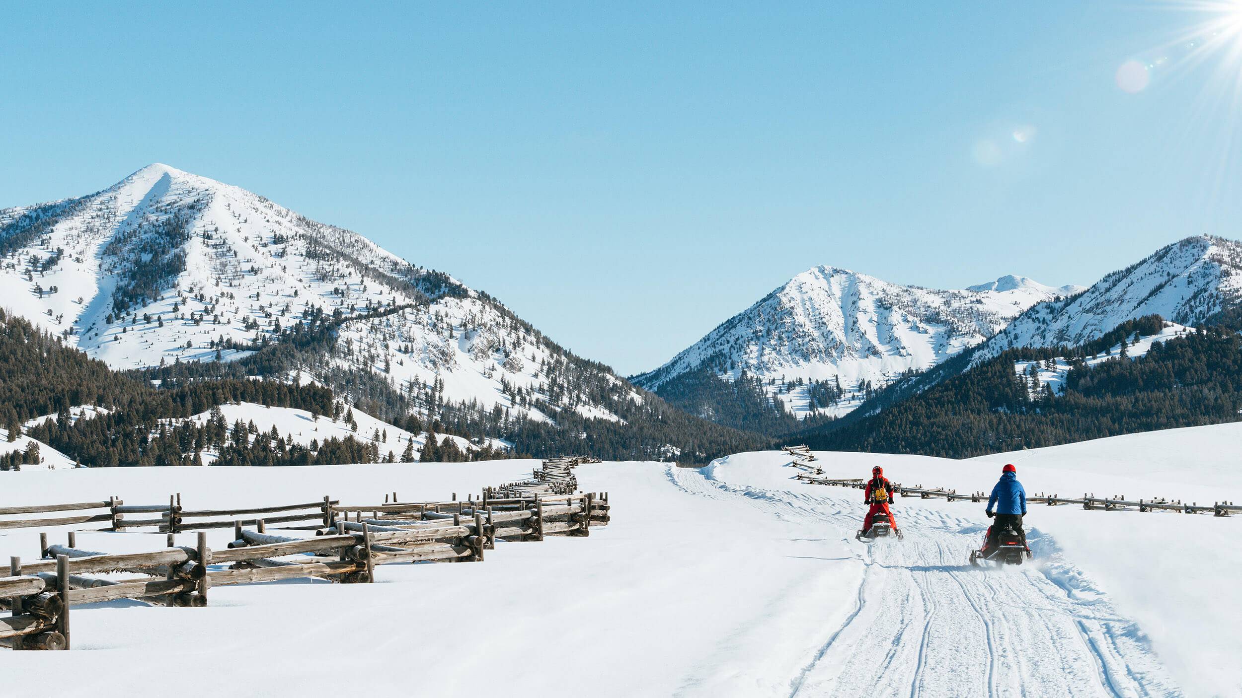 One person in red and another in blue ride snowmobiles through Smiley