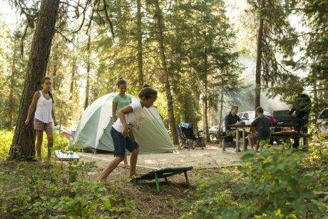 Three children play around their campsite, with tall trees and a sage green colored tent, and white pick up truck in the background.