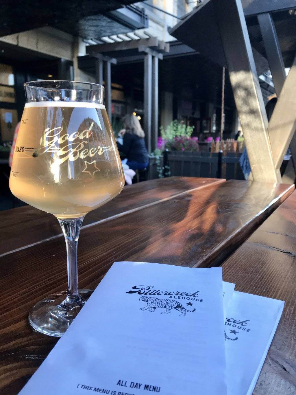 a glass of cider and two menus on a wooden table with people seated at tables in the background