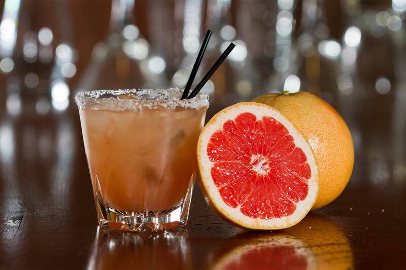 orange colored cocktail with two straws and half a grapefruit leaning against the glass, and a whole grapefruit in the background