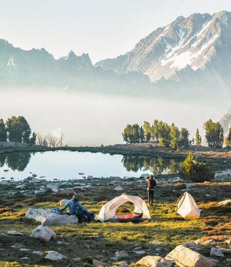 two people at a lakeside campsite with mountains in the distance