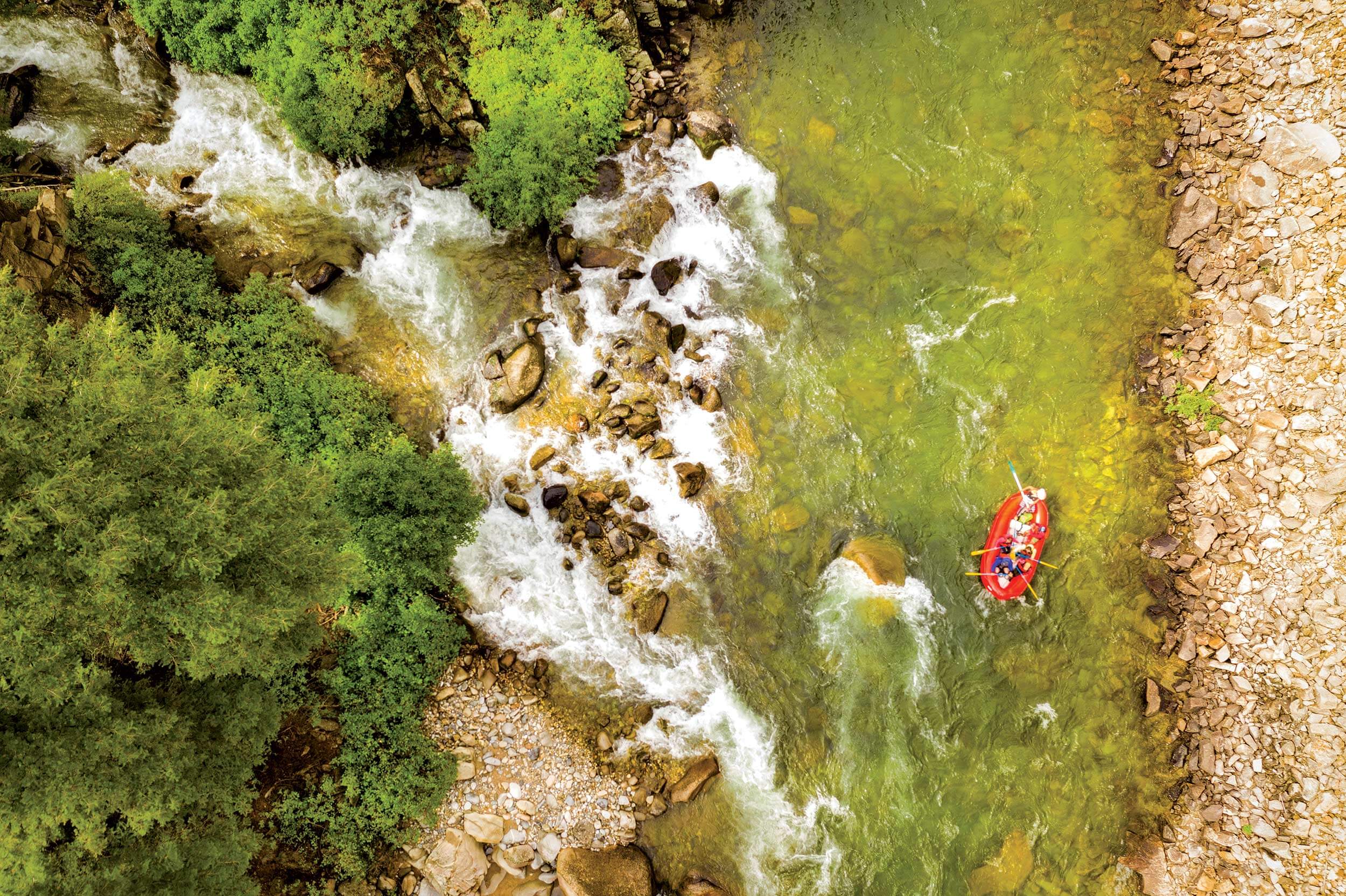 An aerial view of people whitewater rafting, paddling on an orange raft across the Main Salmon River as rapids rush around rocks on waters lined with trees.