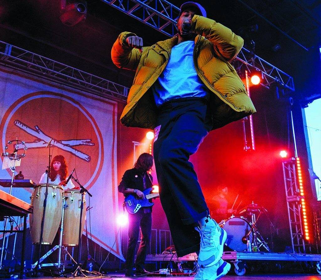 a man singing and dancing on a stage with a guitar play and two drum players in the background