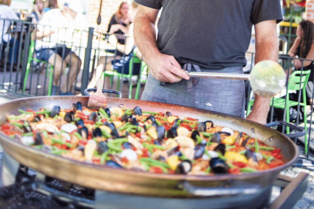 man cooking paella outside and people sitting at tables in the background