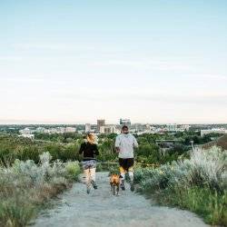 A man and woman running with their dog on a trail lined with tall grass at Camel's Back Park and Boise in the distance.