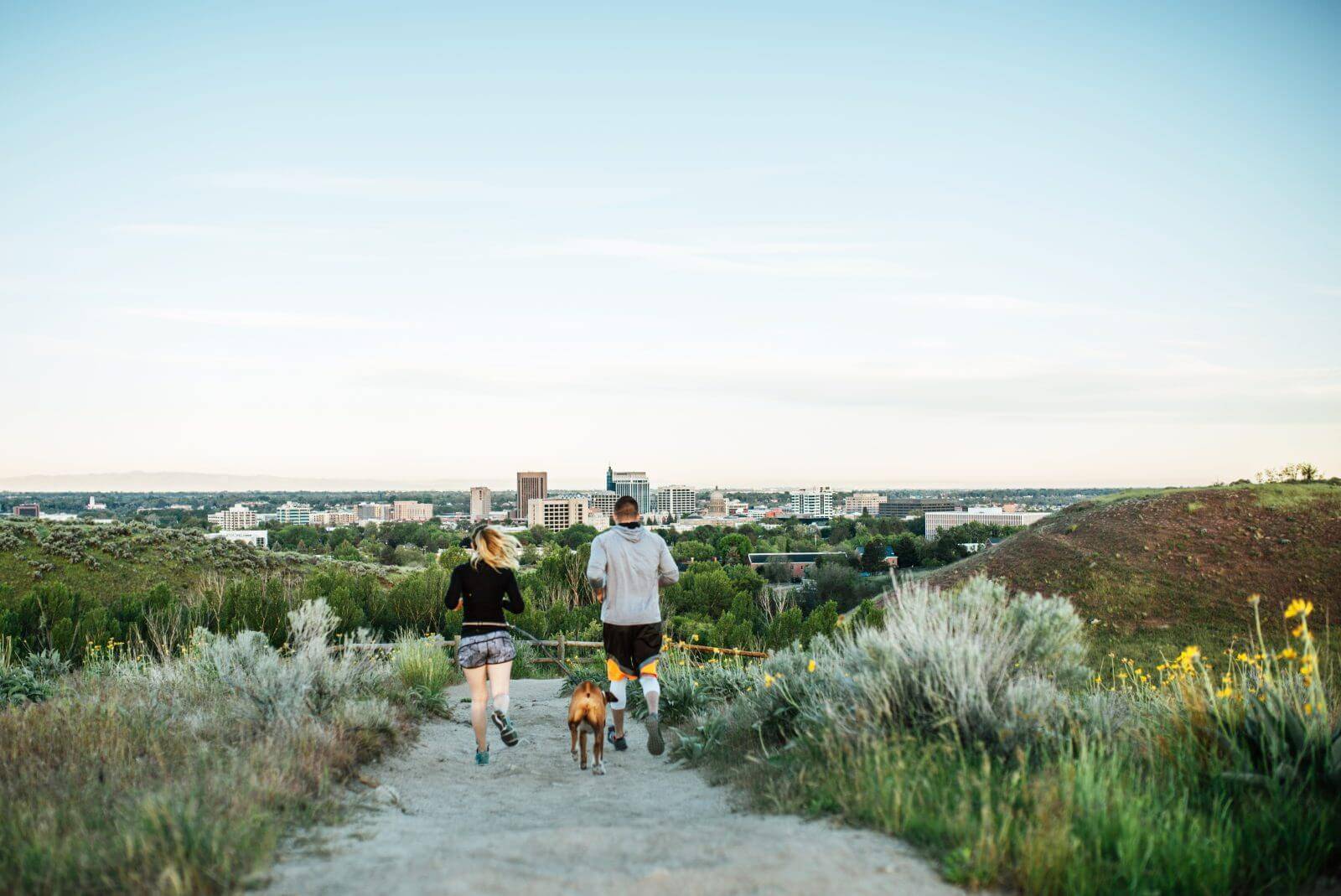 A man and woman running with their dog on a trail lined with tall grass at Camel's Back Park and Boise in the distance.