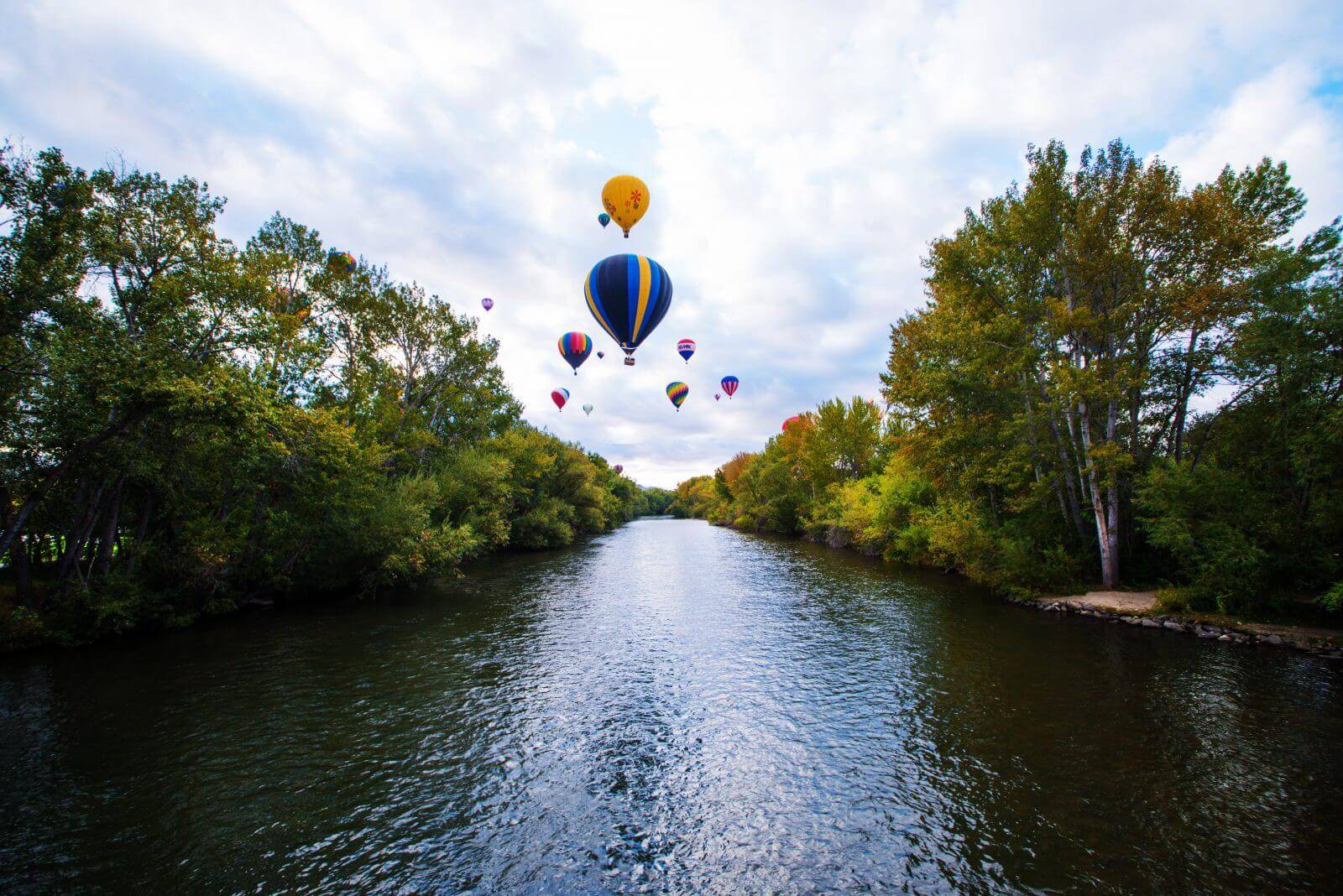 A group of colorful hot air balloons floating above the tree-lined Boise River.