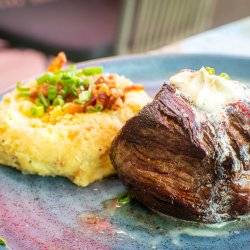 A steak sits at the center of a blueish plate with a serving of garnished mashed potatoes off to the side.