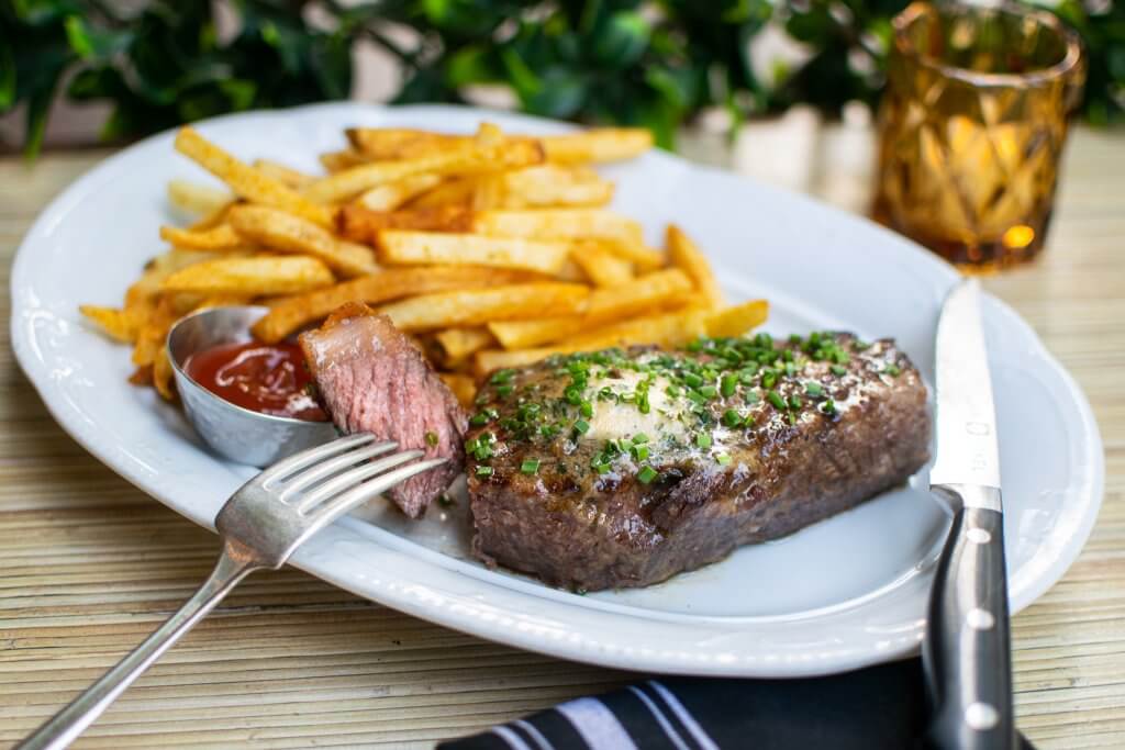 Un filete cubierto de cebolla verde se encuentra en un plato ovalado junto a una porción de papas fritas y una cucharada de ketchup, con un tenedor y un cuchillo en primer plano.