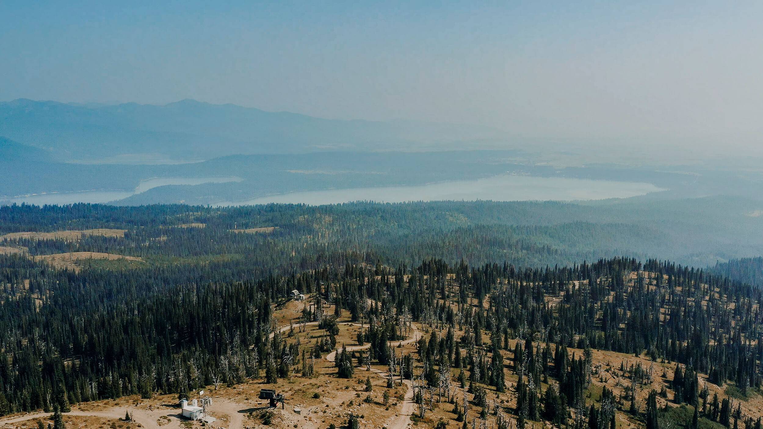 a mountain top view of a forest of trees with a lake and mountains in the distance