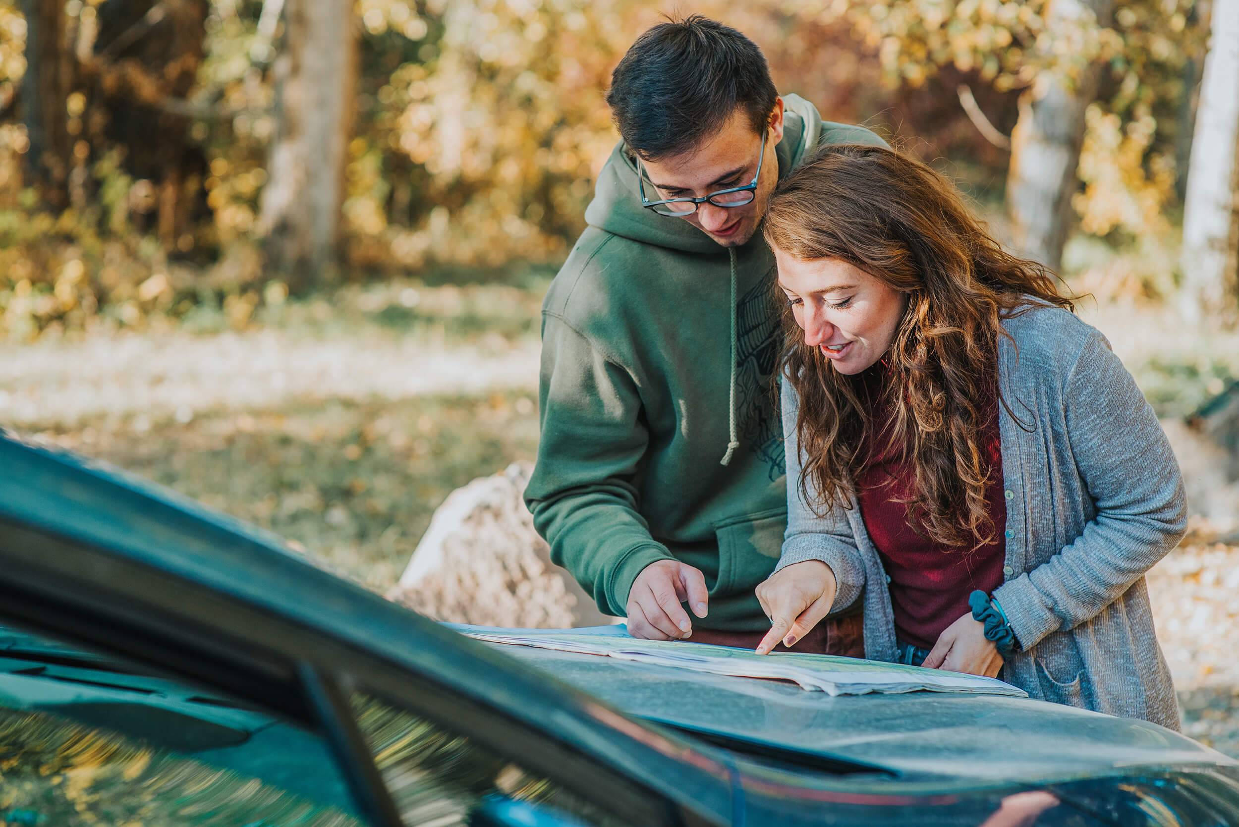 a couple reading a map that's spread out on the hood of a car