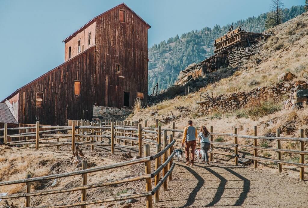 a couple walking with their two dogs along a dirt road towards a tall historic wooden building