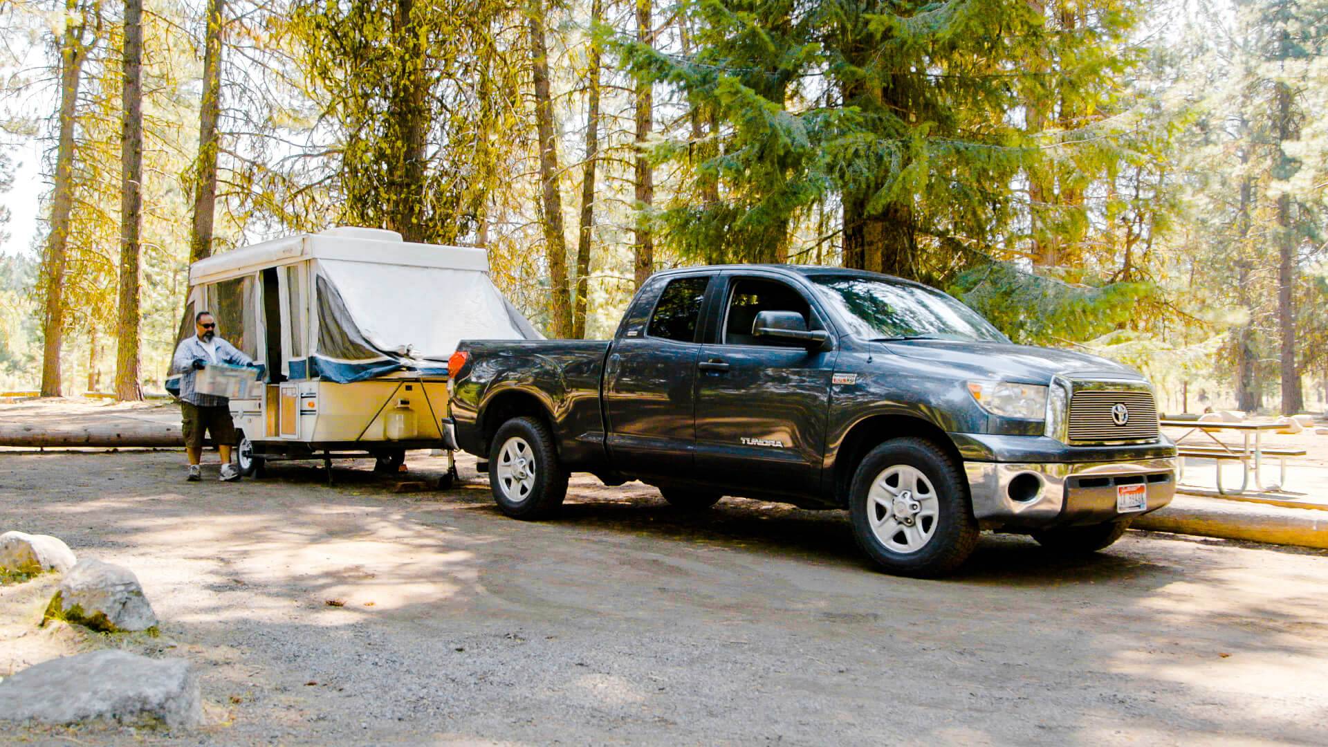 a man carrying a storage container beside a camper hooked up to a toyota tundra in a forest