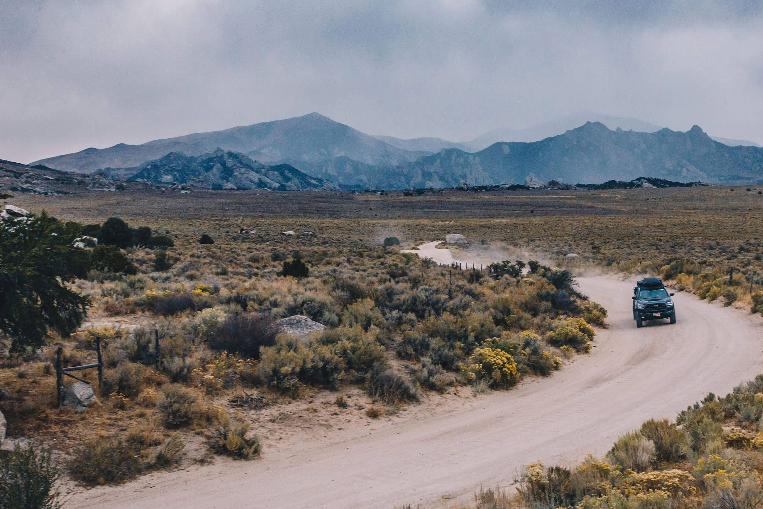 a car driving along a winding dirt road surrounded by desert shrubs with mountains in the background