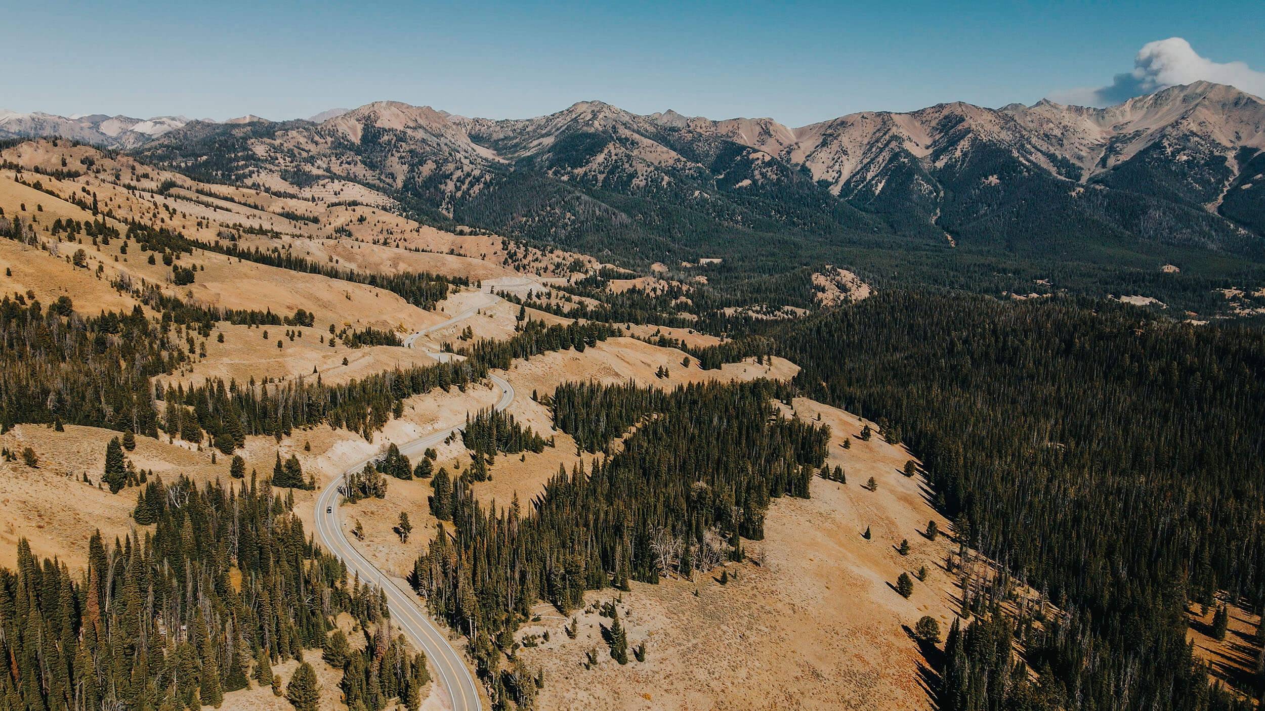 an overhead view of a highway snaking through a tree covered landscape with mountains in the background