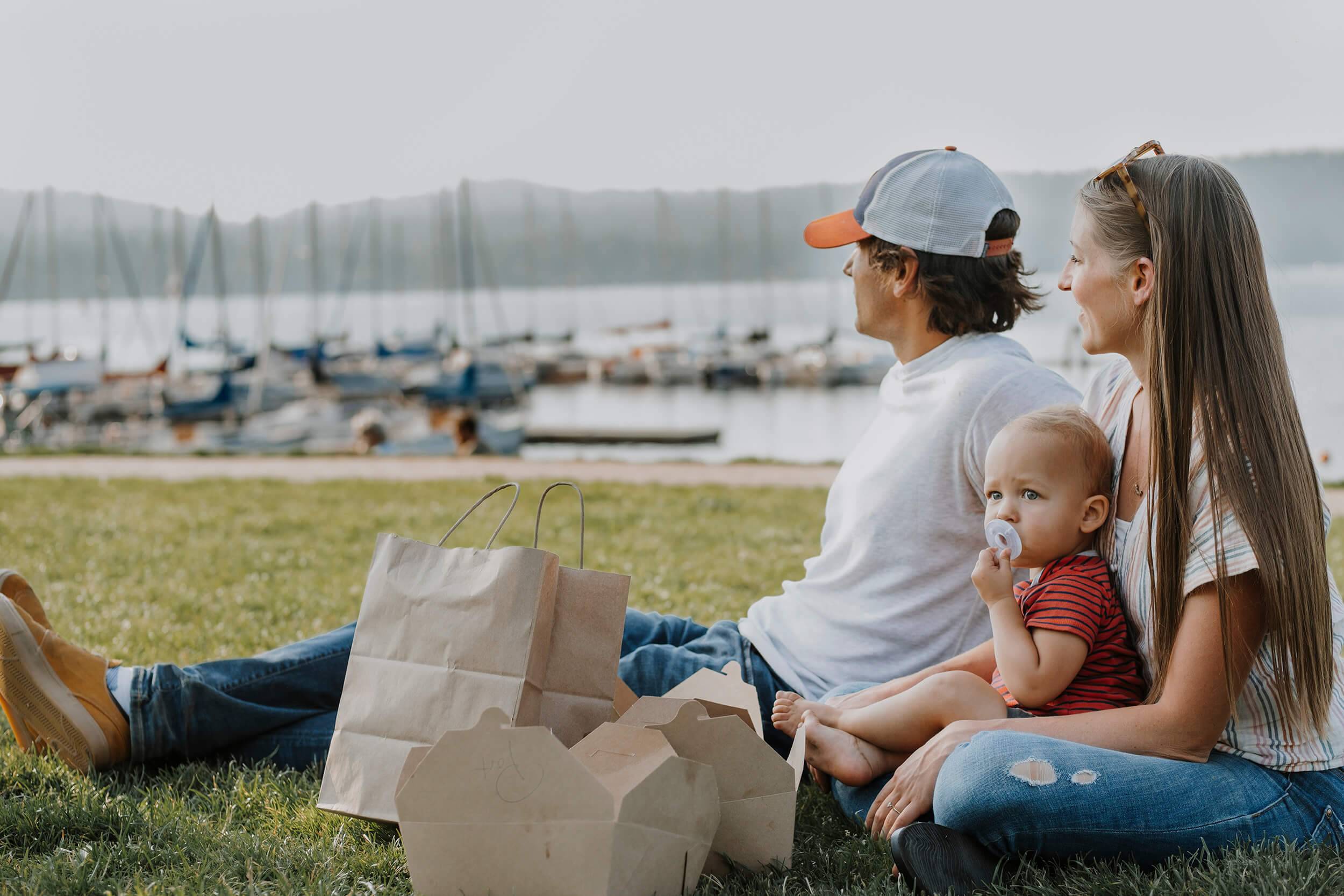 a couple with a baby sitting in the grass with some boats on a lake in the background