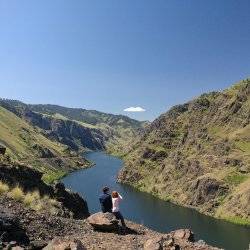 two people sitting on a rock looking out over a river running through a canyon