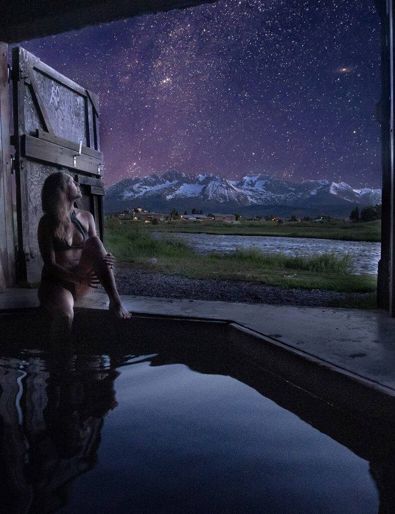 a woman sitting on the edge of a hot spring looking up at a starry sky with a lake and snow-covered mountains in the distance